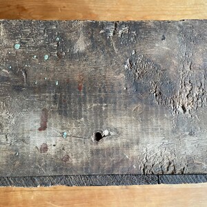 May include: A weathered wooden box with a dark brown finish. The box has a small hole in the center and shows signs of wear and tear, including paint splatters and scratches.