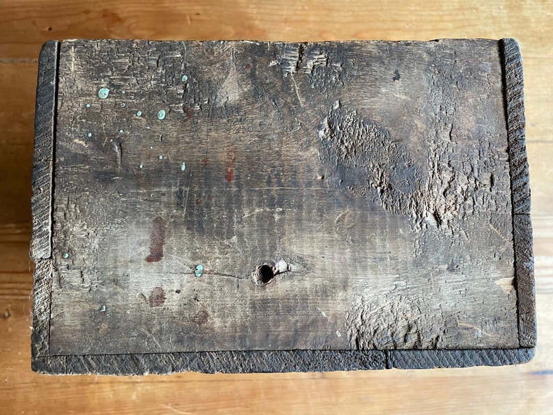 May include: A weathered wooden box with a dark brown finish. The box has a small hole in the center and shows signs of wear and tear, including paint splatters and scratches.