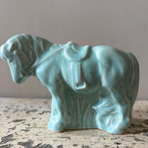 Può includere: Una statuetta di cavallo in ceramica azzurro chiaro con sella e briglia modellate. Il cavallo è raffigurato in posizione eretta con criniera e coda testurizzate, rifinito con uno smalto lucido che mette in risalto i dettagli scolpiti.