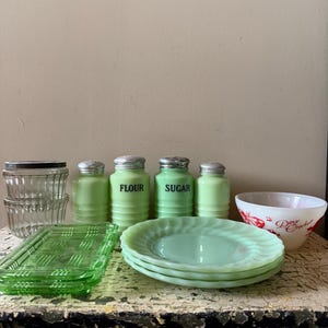 Könnte beinhalten: Eine Sammlung von Vintage-Küchenutensilien in Grün- und Klarglas. Dazu gehören transparente Glasbehälter, grüne Salz- und Pfefferstreuer, Mehl- und Zuckerdosen, Teller, ein Serviertablett und eine Schüssel mit rotem Text.