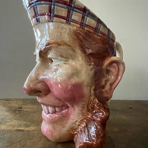 Puede incluir: Un jarro de cerámica con forma de cabeza de hombre con un sombrero a cuadros. El hombre tiene una barba marrón y una amplia sonrisa. El jarro está esmaltado en marrón, rojo y blanco.
