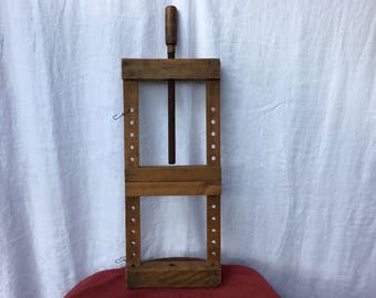 May include: A vintage wooden book press with a threaded rod and a wooden handle. The press has a rectangular frame with multiple holes and metal hooks. The wood has a warm, natural tone.