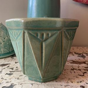 May include: A turquoise ceramic planter with geometric patterns. The planter has a faceted design with vertical and angled lines, creating a textured surface. The planter is a light teal colour and has a vintage aesthetic.