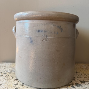 Op de afbeelding: Een vintage steengoed kruik met een lichtbeige afwerking. De kruik heeft een afgeronde body, een licht uitlopende rand en twee kleine handvatten. De tekst "FORT EDWARD, N.Y." en het nummer "3" zijn blauw gedrukt. De kruik wordt waarschijnlijk gebruikt voor voedselopslag.