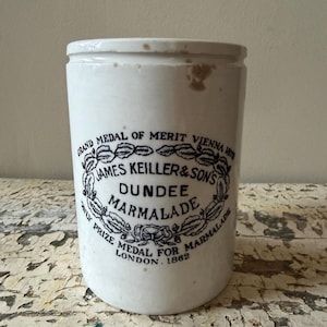 May include: A vintage white ceramic jar with a black and white label. The label reads "James Keiller & Sons Dundee Marmalade" and includes text about awards from Vienna and London. The jar has a cylindrical shape.