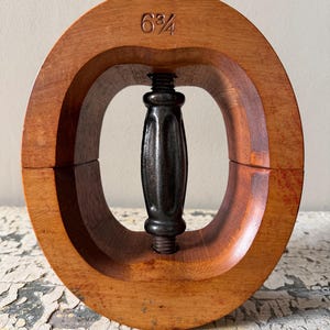 May include: A vintage wooden hat stretcher with a dark metal screw mechanism. The wooden frame is a warm brown color, and the size "6 3/4" is visible on the top. The stretcher is designed to maintain or adjust the size of a hat.