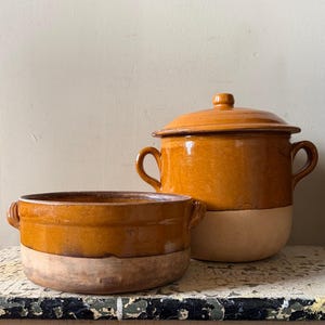 Puede incluir: Una olla alta con tapa y un cuenco ancho y poco profundo, ambos con un diseño bicolor que presenta una sección superior esmaltada en color marrón miel brillante y una base beige mate sin esmaltar. Cada pieza tiene dos pequeñas asas laterales.