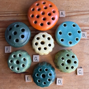 May include: A collection of ceramic flower frogs in various colors, including orange, teal, and green. Each frog has multiple holes for arranging flower stems. The frogs are arranged on a wooden surface, showcasing their different shapes and sizes.