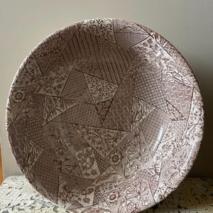 May include: A large, round serving bowl with a patchwork design. The bowl features a variety of brown and cream-colored patterns, including floral, geometric, and textured elements. The bowl has a wide rim and a glossy finish.