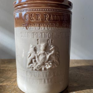 May include: A cylindrical stoneware jar with a brown top and rim, transitioning to a cream-colored body. The jar features embossed text and a crest, with the text "G & E. PAINTER PURVEYORS to HRH THE PRINCE of WALES".