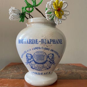 Puede incluir: Un jarrón de cerámica crema con texto azul y un emblema decorativo, que contiene flores de cuentas blancas, verdes y amarillas. El jarrón dice "Moutarde-Diaphane" y "Bordeaux". El jarrón está sobre un libro encuadernado en cuero.
