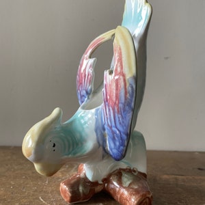 May include: A ceramic bird figurine with a colorful design. The bird has a light yellow head, a turquoise body, and wings with red, blue, and yellow accents. The figurine is perched on a brown base and has a glossy finish.