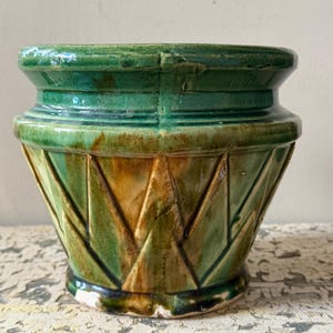 May include: A glazed ceramic planter featuring a vibrant green and amber colour palette. The exterior displays a geometric relief pattern of vertical triangular shapes around the body, with a wide, flared rim and a circular base.