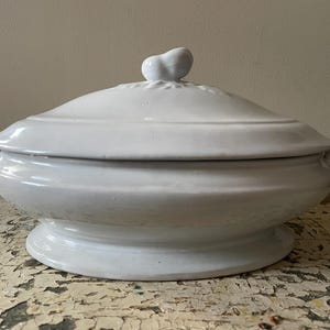 May include: A white ceramic serving dish with a lid. The dish has a round base and a lid with a small, decorative handle. The dish has two small handles on the sides. The dish is likely used for serving food.