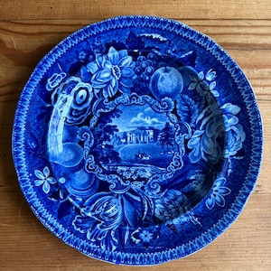 May include: A round, blue and white decorative plate with a detailed floral and landscape design. The plate features a central scene of a building and surrounding landscape, framed by flowers, fruit, and foliage. The rim has a textured, scalloped edge.