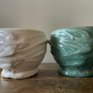Op de afbeelding: Twee keramische kommen met een textuur. Eén is gebroken wit, de andere is turquoise. De kommen hebben een unieke, organische vorm en staan naast elkaar. De kommen zijn waarschijnlijk handgemaakt en geschikt om voedsel in te serveren.