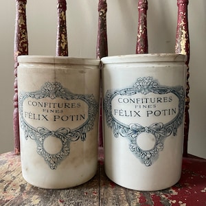 May include: Two vintage white ceramic jars with lids, adorned with elegant blue and white labels reading "CONFITURES FINES FÉLIX POTIN." The cylindrical jars are displayed on a distressed red wooden surface.