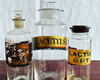 May include: Three vintage glass bottles with labels. The centre bottle has a gold label that reads "ACETYLSA". The bottle on the left has a partially obscured label that reads "USIN COM?. The bottle on the right has a gold label that reads "LACTUC OPT."
