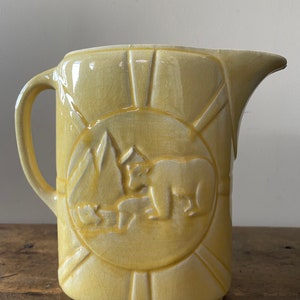 May include: A yellow ceramic pitcher with a raised design of a bear and cubs in a mountain scene. The pitcher has a textured surface and a wide, rounded spout.