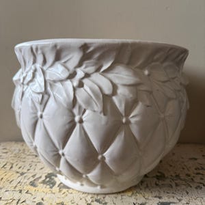 Könnte beinhalten: Ein weißer Keramik-Pflanztopf mit einem Steppmuster und floralen Details am Rand. Der runde Topf hat eine weite Öffnung und ist für den Innen- und Außenbereich geeignet. Der Topf ist aus Keramik.