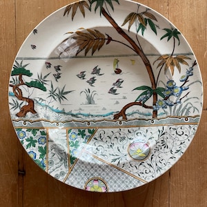 May include: A white ceramic plate with a decorative design featuring a stylized tree with green leaves, brown branches, and yellow flowers. The plate also has a border of green, blue, and yellow floral patterns.