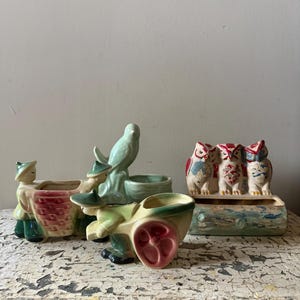 Könnte beinhalten: Eine Sammlung von Vintage-Keramik-Pflanzgefäßen. Die Pflanzgefäße umfassen eine Figur mit einem Korb, einen Vogel mit einem Wagen und drei Eulen, die auf einem Baumstamm sitzen. Die Pflanzgefäße sind in verschiedenen Farben bemalt, darunter Grün, Rot und Creme.