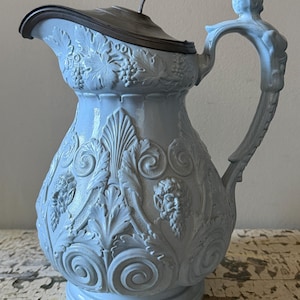 Può includere: Brocca in ceramica azzurra con coperchio e manico color argento. La brocca presenta dettagliate incisioni a rilievo di viti, foglie e figure classiche. Il manico è scolpito con una figura umana.