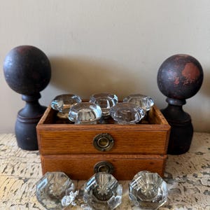May include: A collection of clear, faceted glass knobs and two dark wooden finials. The knobs are displayed in a small wooden box with brass hardware. The finials have a dark finish with red accents. The knobs are a vintage style.