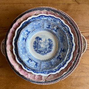 Può includere: Una pila di piatti decorativi antichi. I piatti presentano disegni intricati in blu, rosa e marrone. Il piatto superiore ha una scena paesaggistica blu e bianca. I piatti sono disposti su una superficie di legno.