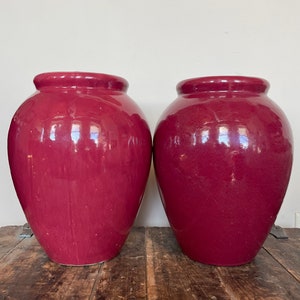 Pode incluir: Dois vasos grandes de cerâmica de cor vermelho escuro com um acabamento brilhante. Os vasos são redondos e têm uma abertura larga na parte superior.