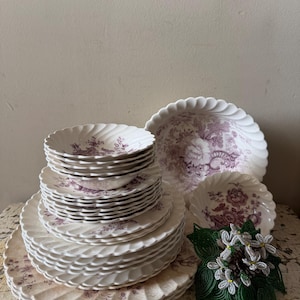 May include: A stack of cream-colored ceramic plates and bowls featuring a purple floral transferware pattern and scalloped edges. A separate larger bowl and a smaller bowl with matching patterns are placed nearby, alongside a beaded floral ornament.