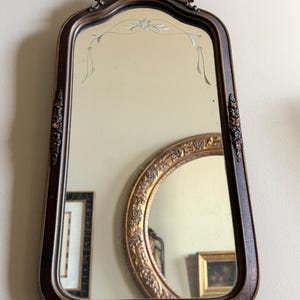 Może przedstawiać: Antyczne lustro w brązowej ramie z ozdobnym topem i kwiatowymi akcentami. Lustro ma dekoracyjny, wytrawiony wzór na górze. Złote lustro w ramie odbija się w szkle.