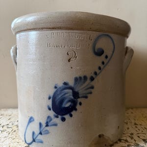 Pode incluir: Um pote de grés cilíndrico com acabamento em tom bege claro, apresentando duas alças laterais e um motivo floral pintado em azul. A frente está estampada com o texto S. B. MOWBRAY HAMMOND 2, indicando o fabricante e o tamanho 2.