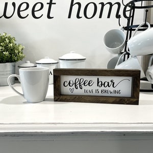 May include: A wooden sign with the text "coffee bar" and the phrase "love is brewing" in cursive script. The sign is brown with a white background and a heart symbol. The sign is sitting on a white counter with a white coffee mug and other kitchen items.