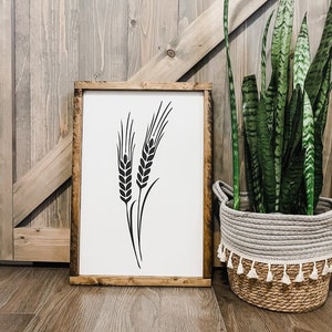 May include: A framed print of a black wheat stalk design on a white background. The frame is made of wood with a brown stain. The print is sitting on a wooden floor next to a potted plant.