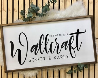 May include: A white wooden sign with a brown frame. The sign says "Wallcraft" in black cursive lettering with "EST. 08.10.2019" above the name and "SCOTT & KARLY" below.