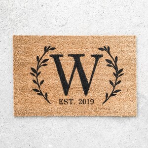 May include: A brown coir doormat with a black wreath design and the letter "W" in the centre. The text "EST. 2019" is printed below the wreath.