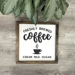 May include: A white wooden sign with black text that reads "Freshly Brewed Coffee" with a coffee cup icon and "Cream - Milk - Sugar" below. The sign is framed in dark wood and is sitting on a light wood surface.