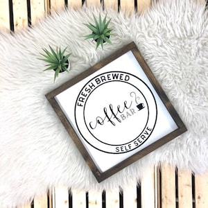May include: A white wooden sign with a brown frame, featuring a black circle with the text "Fresh Brewed Coffee Bar Self Serve" and a stylised coffee cup icon.