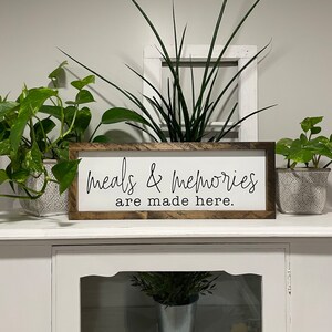 May include: A wooden sign with the text "meals & memories are made here." The sign is brown with a white background and black lettering. The sign is sitting on a white shelf with plants in pots on either side.