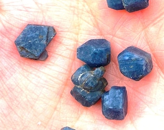 May include: A collection of six dark blue sapphire crystals, some with a hexagonal shape, others with a more rounded shape.