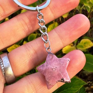 May include: A pink star-shaped gemstone keyring with a silver chain. The gemstone has a textured surface and is suspended by a silver chain. A unique accessory.