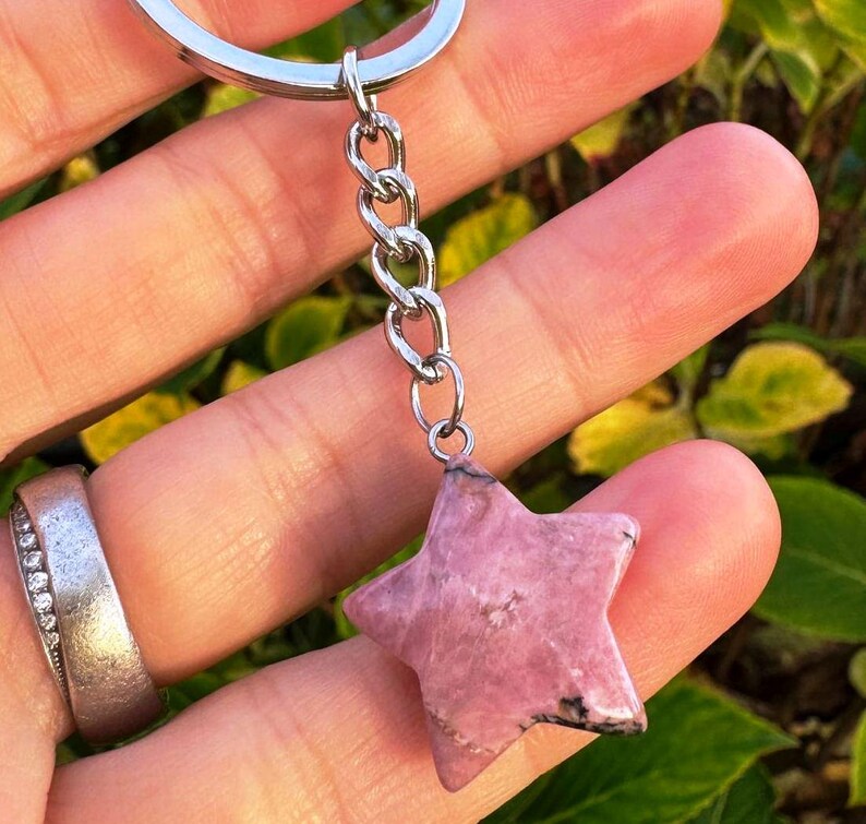 May include: A pink star-shaped gemstone keyring with a silver chain. The gemstone has a textured surface and is suspended by a silver chain. A unique accessory.