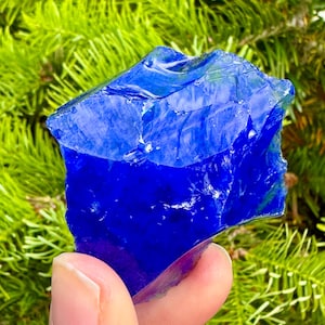 May include: A vibrant, deep blue glass rock with a jagged, irregular shape. The translucent stone is held against a backdrop of green foliage, highlighting its rich color and texture. The glass appears to have a glossy finish.