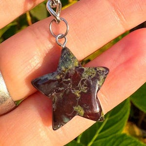May include: A star-shaped pendant with a silver chain. The gemstone has a dark maroon base with green and yellow veining. The pendant is held up by a silver ring and chain. The background is a blurred view of a hand and green foliage.