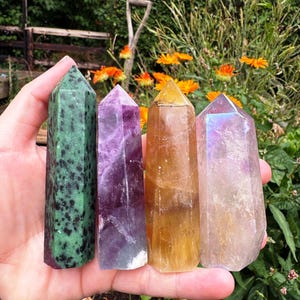 Può includere: Quattro torri di cristallo lucido nei toni del verde, viola, giallo e iridescente. I cristalli sono tenuti in mano su uno sfondo di fiori arancioni e verde. Ogni cristallo ha una parte superiore appuntita.