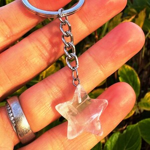 May include: A clear quartz star-shaped keyring charm. The charm hangs from a silver chain and keyring. The star is translucent with internal patterns. The background is a blurred mix of green and yellow.