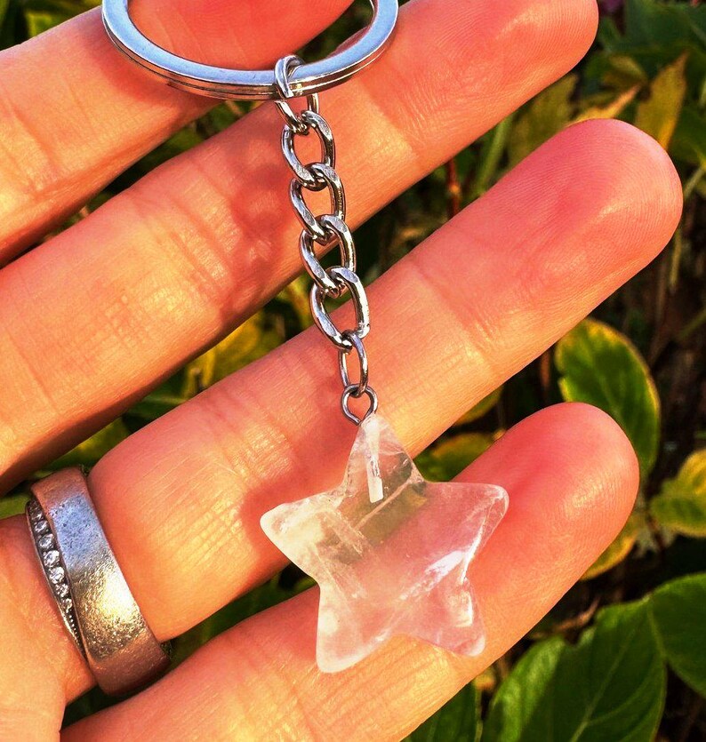 May include: A clear quartz star-shaped keyring charm. The charm hangs from a silver chain and keyring. The star is translucent with internal patterns. The background is a blurred mix of green and yellow.