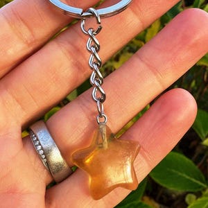 May include: A keyring with a silver chain and a star-shaped charm. The charm is a translucent amber colour. The keyring is held in a hand, with a silver ring visible on a finger.