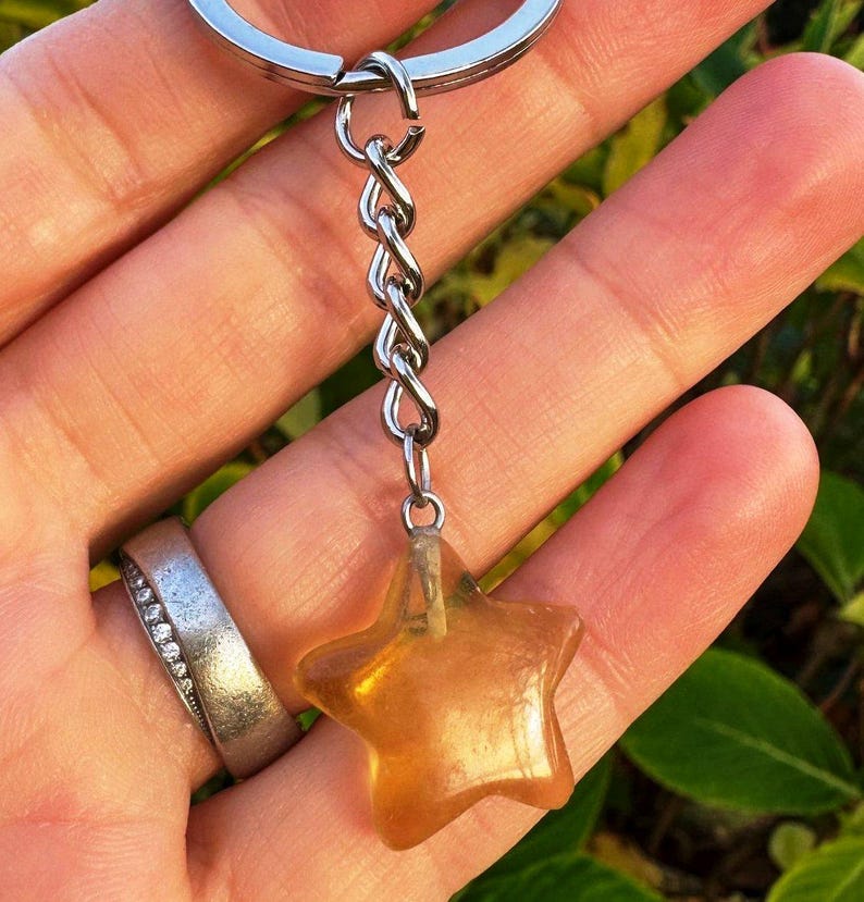 May include: A keyring with a silver chain and a star-shaped charm. The charm is a translucent amber colour. The keyring is held in a hand, with a silver ring visible on a finger.